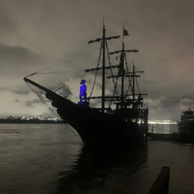 Navio pirata atracado à noite, em silhueta, com as luzes da cidade ao fundo
