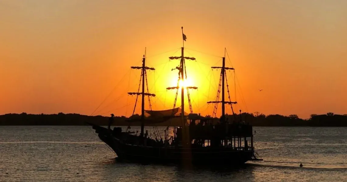 Navio pirata Fantástico em silhueta, com o sol se pondo atrás dos mastros no Guaíba