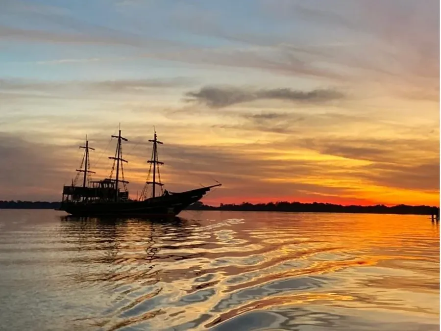 Navio pirata em silhueta no Guaíba durante o pôr do sol, com o céu alaranjado refletido na água
