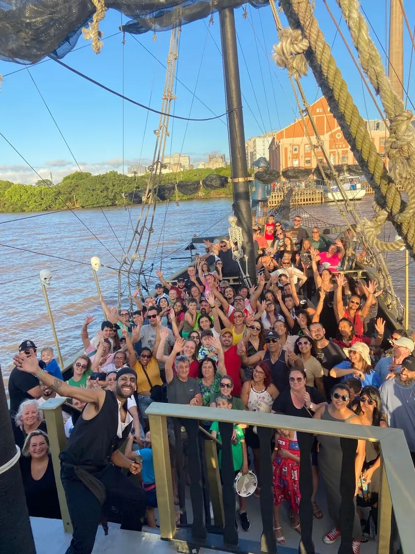 Passageiros acenando no convés do navio pirata, com um pirata da tripulação em primeiro plano e o Guaíba ao fundo
