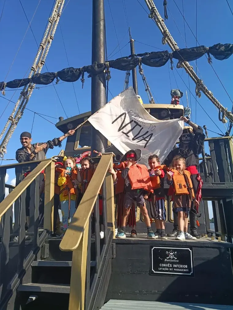 Crianças com coletes salva-vidas laranja no convés do navio pirata, acenando com uma bandeira